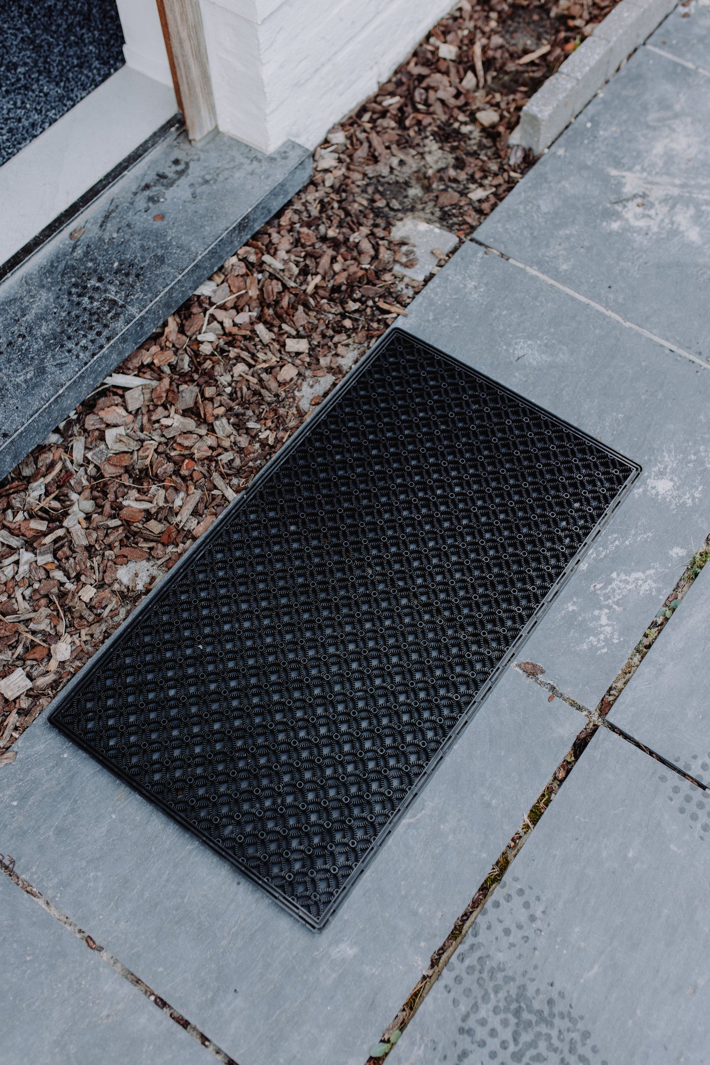 Outdoor black scraper mat with a distinctive pattern, positioned in front of an open door on a grey stone floor.