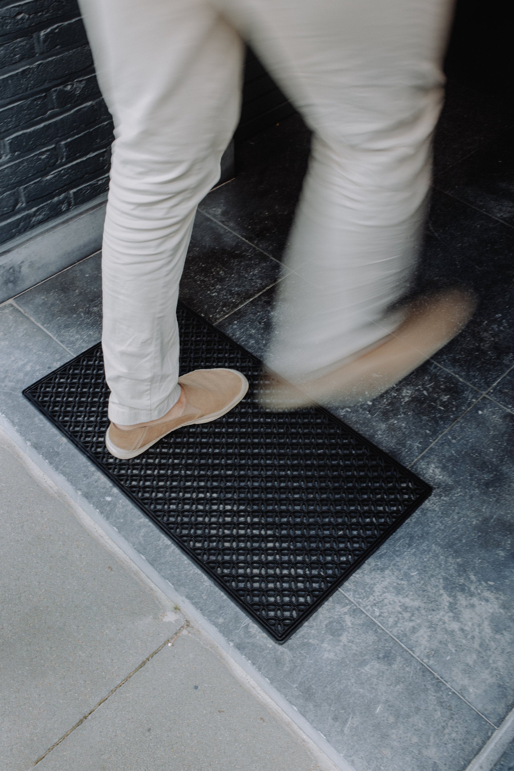 Man wearing brown shoes walking across a black scraper mat while entering a house.