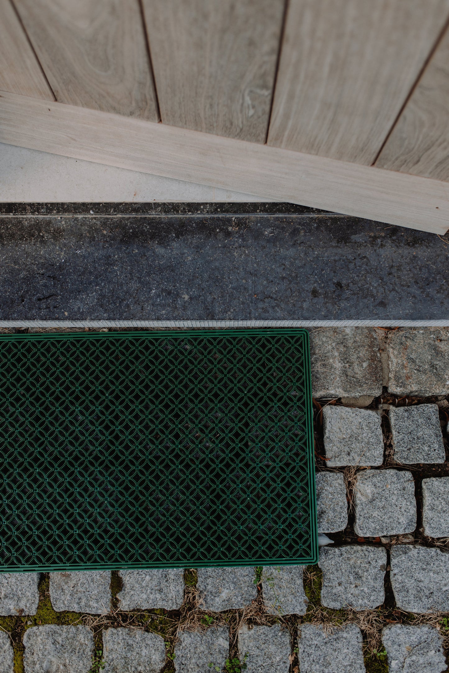 Unique outdoor green scraper mat with an eye-catching pattern, positioned in front of an open door on a grey stone floor