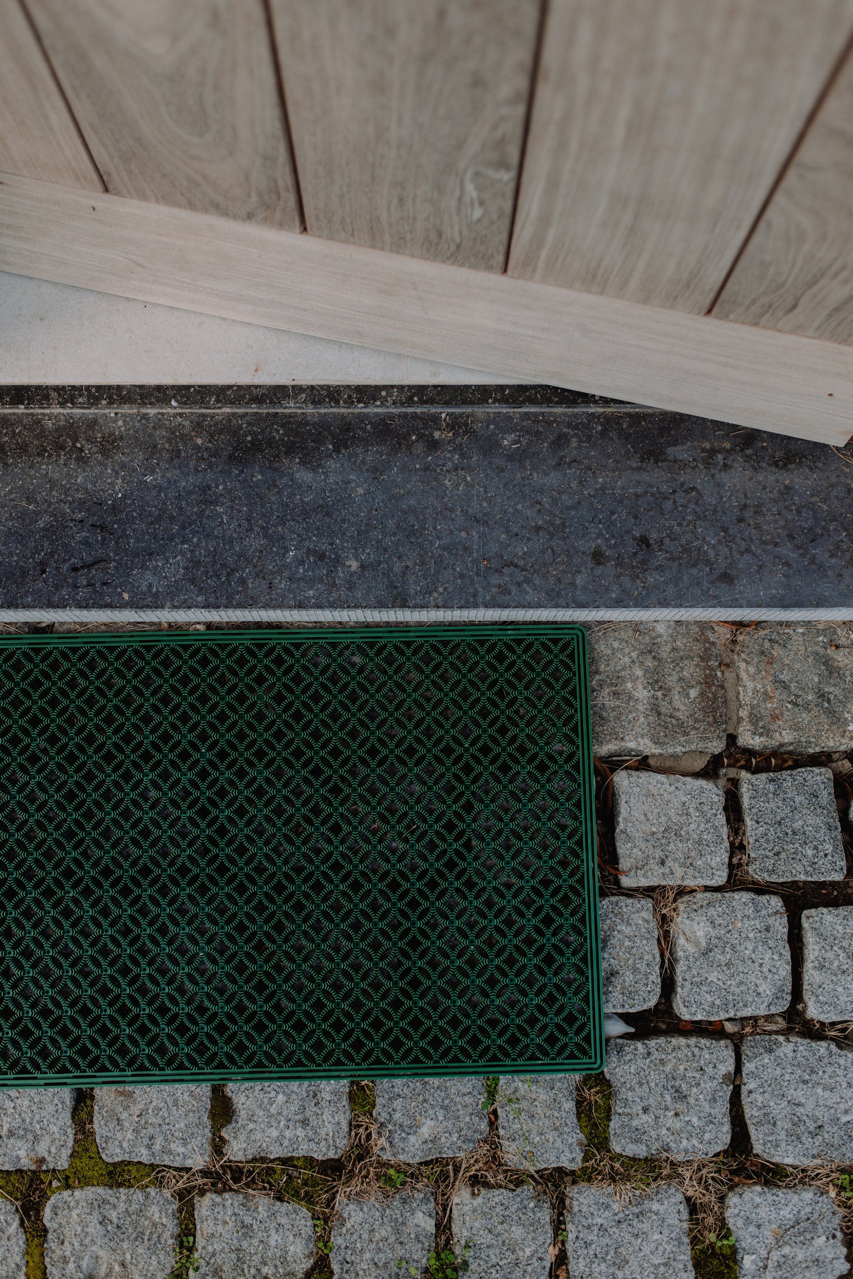 Unique outdoor green scraper mat with an eye-catching pattern, positioned in front of an open door on a grey stone floor